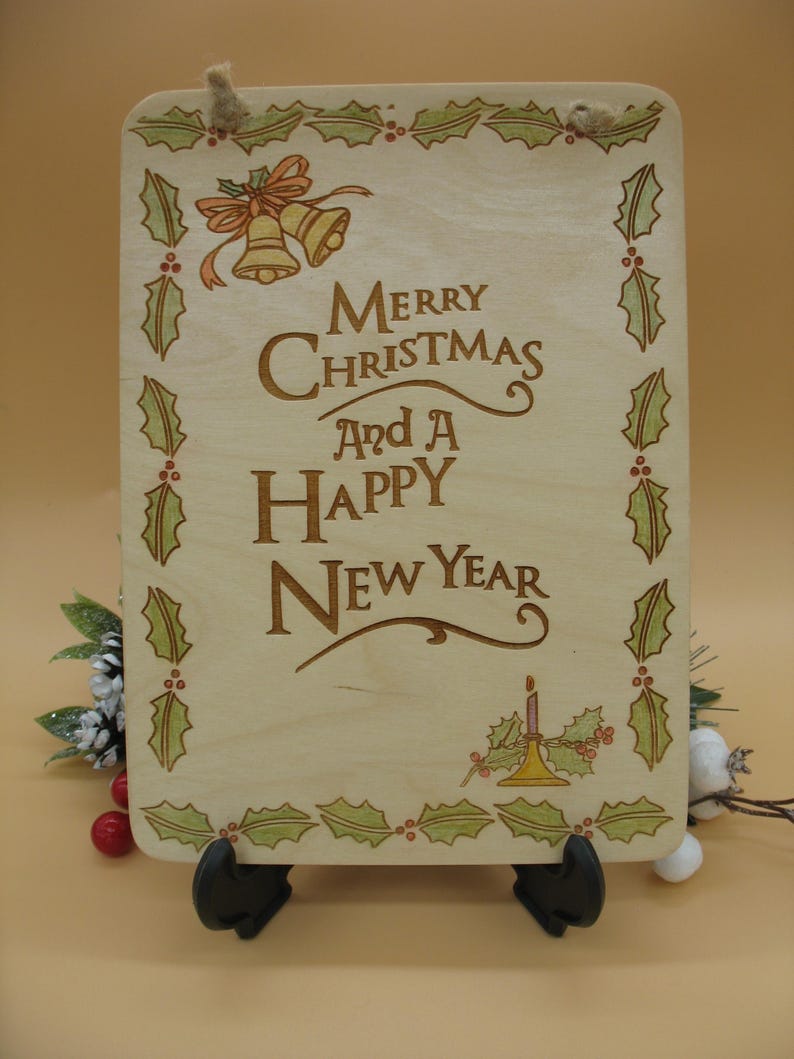 May include: Wooden Christmas sign with the message "Merry Christmas and a Happy New Year" in brown lettering. The sign is decorated with holly leaves, berries, and bells. A jute rope is attached for hanging.