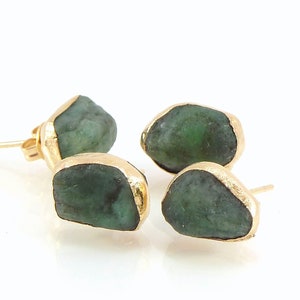 May include: Four gold-plated stud earrings with rough-cut, deep green emerald stones. The emeralds are set in a gold bezel, creating a contrast of textures and colors. The earrings are displayed on a white surface.