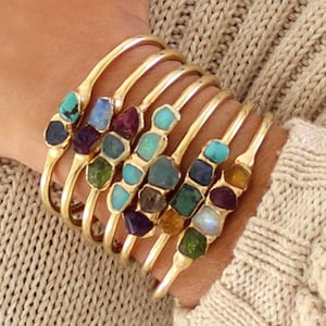May include: A set of gold bangle bracelets with different colored gemstones. Each bracelet features a row of small, irregular shaped gemstones in various colors, including turquoise, amethyst, opal, and emerald.