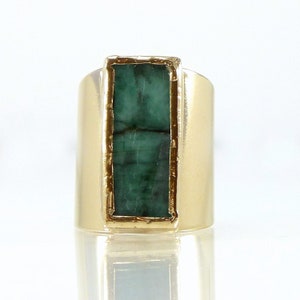 Peut inclure: Une bague en or ornée d'une pierre précieuse rectangulaire vert émeraude. La bague a un large anneau et une monture en or qui encadre l'émeraude. La surface de la pierre précieuse présente des inclusions naturelles visibles. La bague est présentée sur fond blanc.