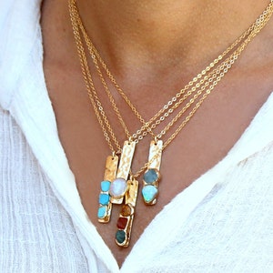 May include: A gold chain necklace with four pendants, each featuring a different gemstone. The pendants are rectangular and have a hammered texture. The gemstones include turquoise, opal, and a pearl.