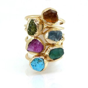 Puede incluir: Una colección de anillos de oro, cada uno con una gema única de forma irregular. Las gemas varían en color, incluyendo tonos de marrón, verde, azul, morado y turquesa. Los anillos se muestran sobre un fondo blanco.