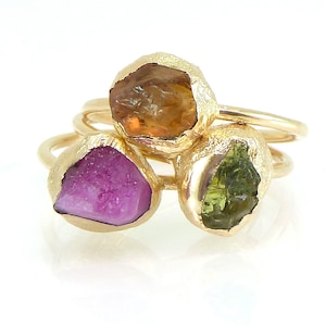 May include: Three gold rings with unique gemstone settings. One ring features a vibrant orange stone, another a deep pink stone, and the third a green stone. The rings are stacked, showcasing the variety of colors and textures.