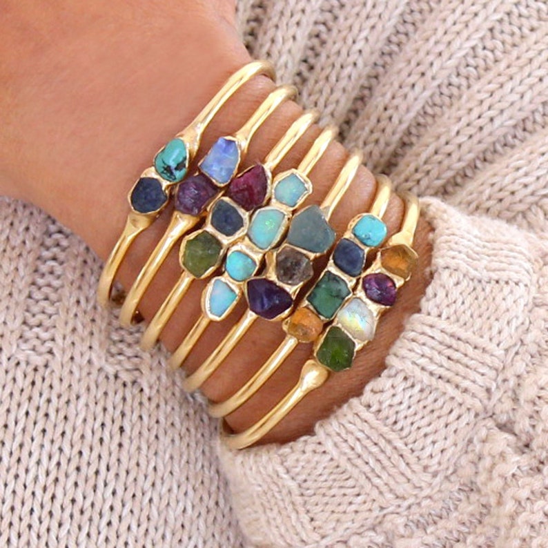 May include: A set of gold bangle bracelets with colorful gemstone accents. Each bracelet features a row of different colored stones, including turquoise, blue, green, and purple.