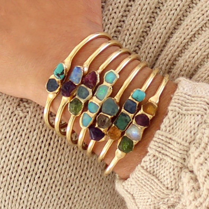 May include: A set of gold bangle bracelets with different colored gemstones. Each bracelet features a row of small, irregular shaped gemstones in various colors, including turquoise, amethyst, opal, and emerald.