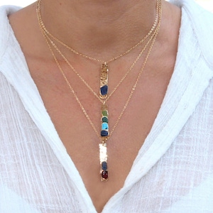 May include: Layered gold necklace with three pendants. The top pendant is a hammered gold bar with a blue stone. The middle pendant is a gold bar with four stones in shades of green, blue, and turquoise. The bottom pendant is a gold bar with a red stone.