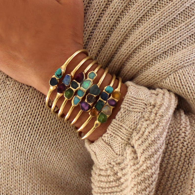 May include: A set of gold bangle bracelets with colorful gemstone accents. Each bracelet features a different gemstone, including turquoise, amethyst, and emerald.