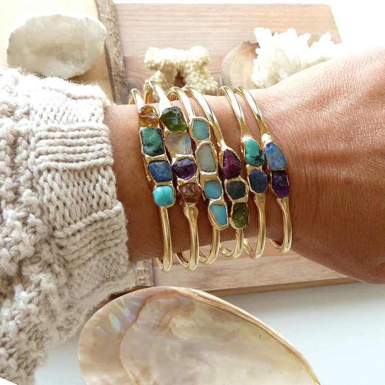 May include: A set of six gold bangles with colorful gemstone accents. Each bangle features a different combination of stones, including turquoise, amethyst, opal, and green stones.