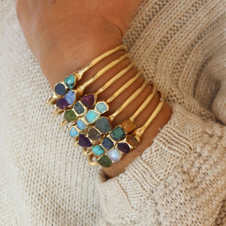 May include: A set of gold bangles with different colored gemstones. The gemstones are set in a cluster on the bangle and are in various shapes and sizes.