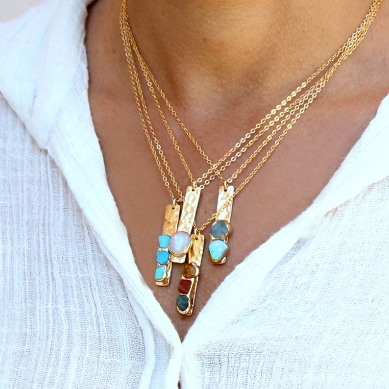 May include: A gold chain necklace with four pendants, each featuring a different gemstone. The pendants are rectangular and have a hammered texture. The gemstones include turquoise, opal, and a pearl.