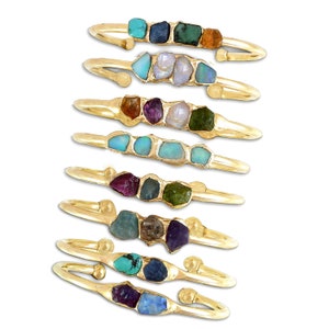 May include: A set of eight gold bangle bracelets, each featuring a row of colorful gemstones. The gemstones are set in a raw, uncut style, and each bracelet has a different combination of colors. The bracelets are stacked on top of each other, creating a colorful and eye-catching display.