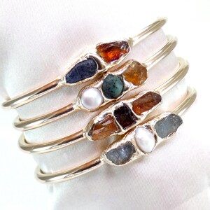 May include: Set of four gold bangles with different colored gemstones and pearls. Each bangle features a unique combination of stones, including brown, blue, green, and white.