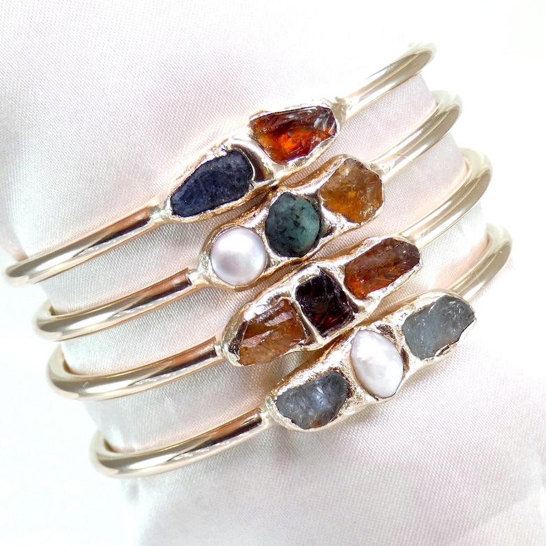 May include: Set of four gold bangles with different colored gemstones and pearls. Each bangle features a unique combination of stones, including brown, blue, green, and white.