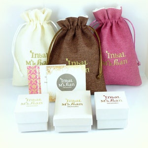 May include: Three white boxes with the text "Inbal MiShan TREASURES" printed in gold. The boxes are sitting on a table in front of three small fabric bags. The bags are beige, brown, and pink and have the text "Inbal MiShan" printed in gold.