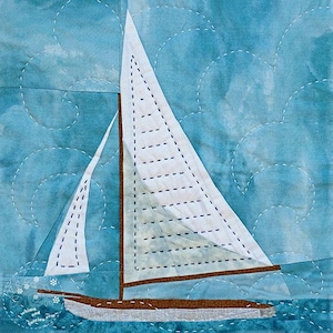 Op de afbeelding: Een gequilt vierkant met een zeilboot op een blauwe achtergrond. De boot heeft een wit zeil met zwarte stiksels en een bruine romp. De achtergrond heeft een water- en hemelontwerp met witte gestikte krullen. De tekst "whims and fancies.com" staat linksonder.