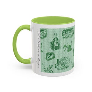 May include: White ceramic mug with a green interior and handle. The exterior features a light green pattern with various green illustrations. The text "Julie Williamson Designs" is printed vertically on the side of the mug.