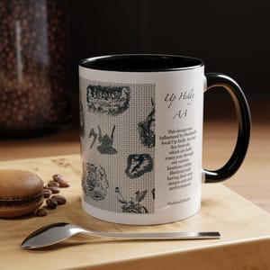 Puede incluir: Taza de cerámica blanca con interior y asa negros. La taza presenta un diseño con varias ilustraciones y el texto "Up Helly AA". Una cuchara y un macaron están sobre una superficie de madera.