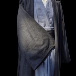 Könnte beinhalten: Ein graues Jedi-Roben- und Tunika-Set, das auf einer Schaufensterpuppe ausgestellt ist. Die Robe hat ein langes Design mit Kapuze und weiten Ärmeln. Die Tunika ist hellblau mit Wickelverschluss. Das Outfit ist für Cosplay oder Kostüme gedacht.