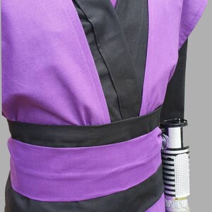 Puede incluir: T&uacute;nica morada y negra con un cintur&oacute;n negro y una empu&ntilde;adura de sable l&aacute;ser plateada unida al lado.
