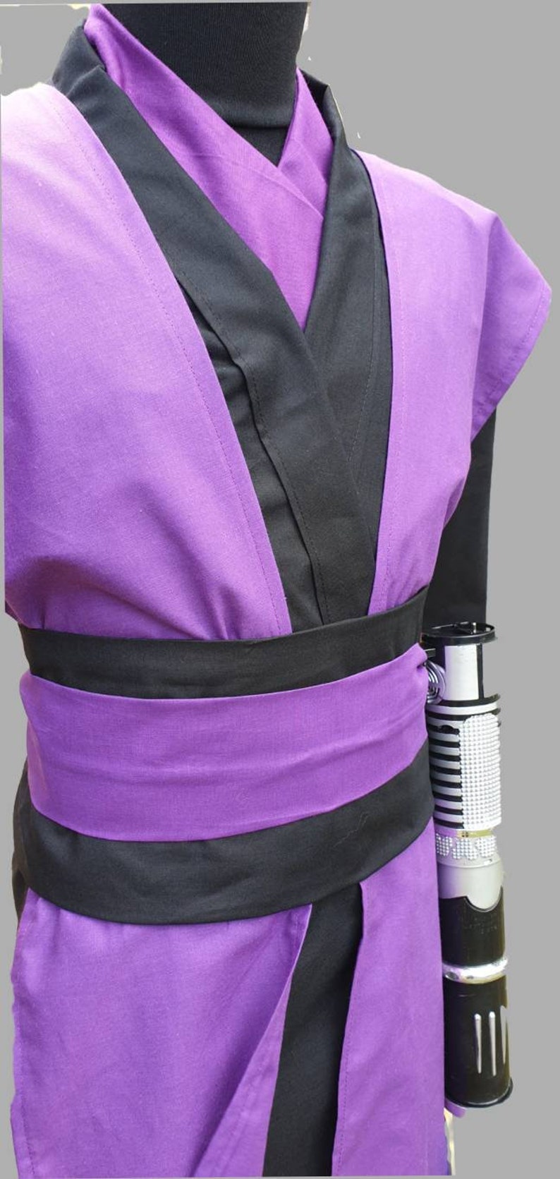 Puede incluir: T&uacute;nica morada y negra con un cintur&oacute;n negro y una empu&ntilde;adura de sable l&aacute;ser plateada unida al lado.