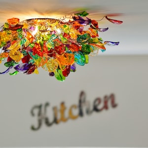 以下が含まれることがあります： さまざまな形とサイズのカラフルなガラス製シャンデリア。シャンデリアは天井から吊り下げられており、点灯しています。背景には「キッチン」という言葉が見えます。