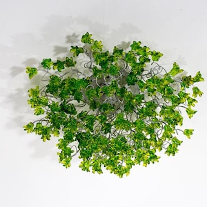 Könnte beinhalten: Draufsicht auf eine dekorative Deckenleuchte. Die Leuchte zeigt eine Ansammlung von grünen und gelben Glasblättern, die an silberfarbenen Metallzweigen befestigt sind. Die Leuchte hängt vor einem weißen Hintergrund.