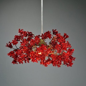 May include: A red floral chandelier with a brass-coloured base and a silver-coloured hanging rod. The chandelier is composed of many red flower-shaped crystals. The light fixture is suspended against a grey background.