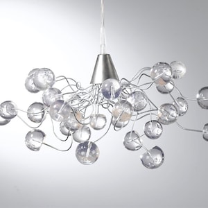 May include: Contemporary chandelier with multiple clear glass spheres suspended from a silver metal frame. The light fitting has a cone-shaped canopy and thin, curved metal arms, creating a unique design. The spheres are various sizes.