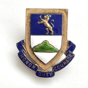 Pode incluir: Um alfinete de metal dourado vintage com um design de escudo. O escudo apresenta uma parte superior azul com um leão dourado, um centro branco e uma montanha verde. As palavras "Honra", "Dever" e "Coragem" são exibidas em azul.