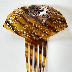 May include: A vintage hair comb with a fan-shaped top and a series of tines. The comb is made of a tortoiseshell material, featuring a warm brown and amber color palette. The top is intricately carved with floral and leaf patterns.