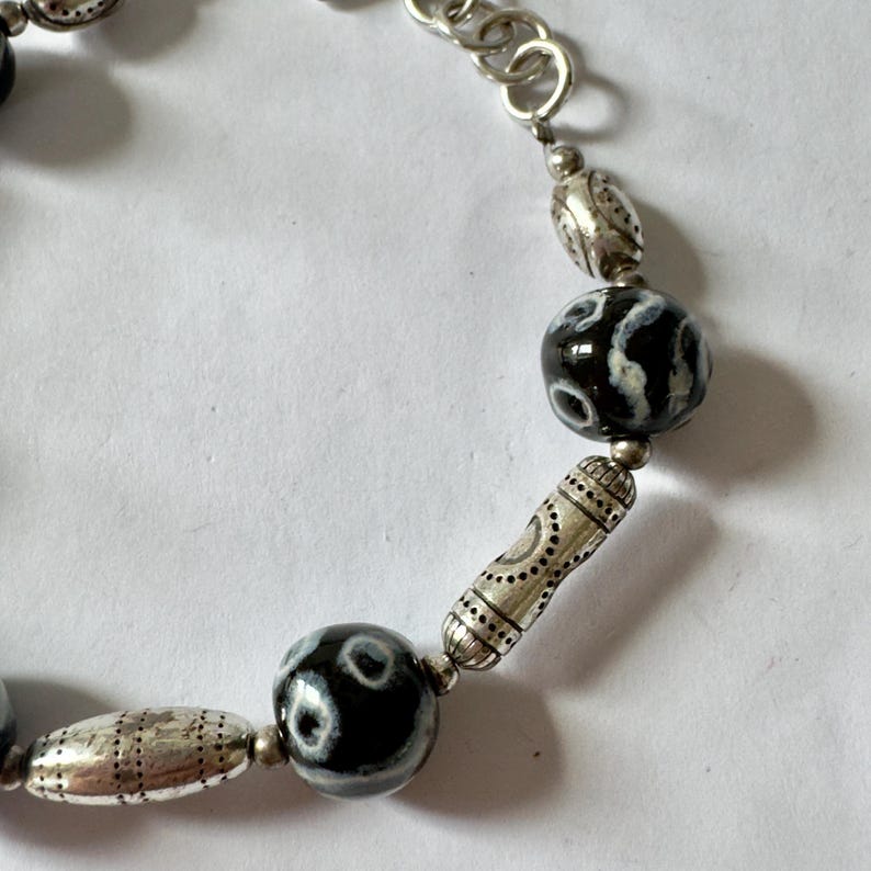 May include: A silver chain bracelet featuring black and white patterned beads and silver-toned metal accents. The bracelet has a mix of round and oval beads, with a toggle clasp. The design is intricate and detailed.