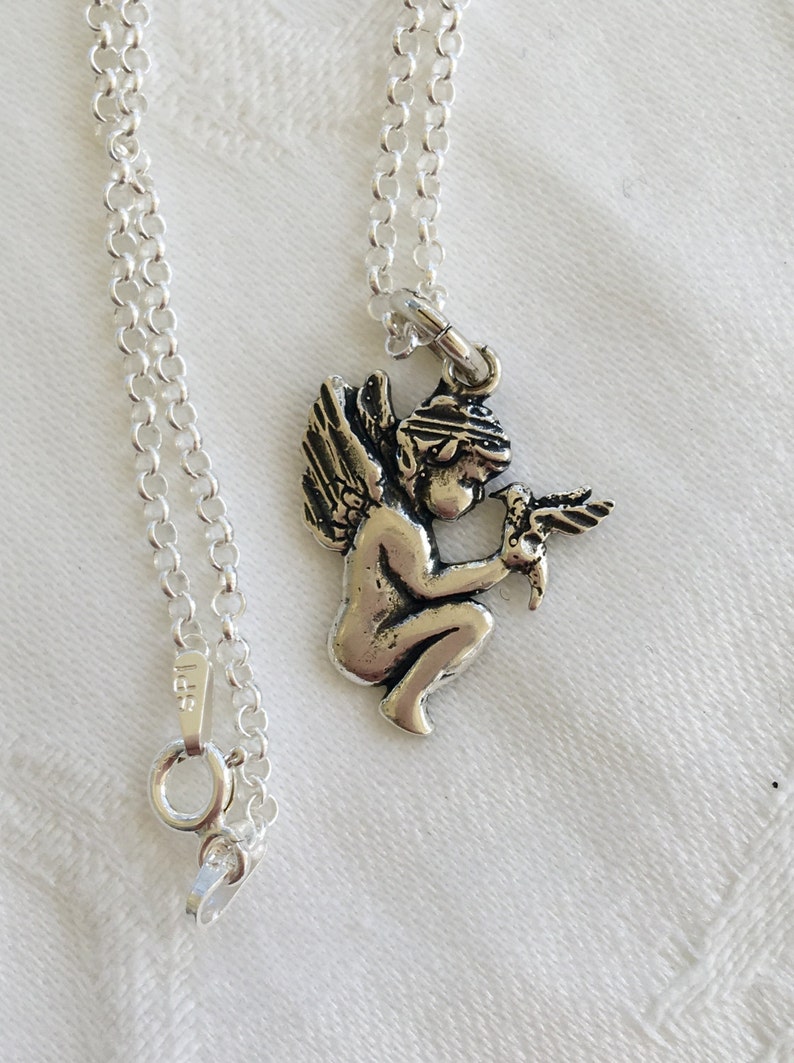 May include: A silver-colored necklace featuring a small pendant of a cherub holding a bird. The pendant is attached to a delicate chain with a lobster clasp. The necklace is displayed on a white surface.