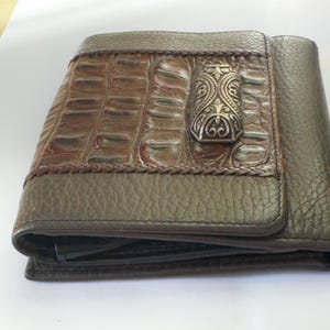 Puede incluir: Una cartera de cuero marr&oacute;n con una solapa con textura de cocodrilo y un cierre decorativo plateado. La cartera tiene un dise&ntilde;o de dos pliegues y un ribete trenzado. El cuero parece ser una combinaci&oacute;n de marr&oacute;n y verde oliva.