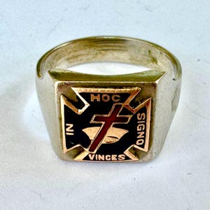 Könnte beinhalten: Ein silberfarbener Ring mit einer quadratischen Vorderseite und einem schwarzen Emaille-Kreuz-Emblem. Das Kreuz ist mit einem roten Akzent versehen und der lateinische Text IN HOC SIGNO VINCES ist in goldfarbenen Buchstaben eingraviert.