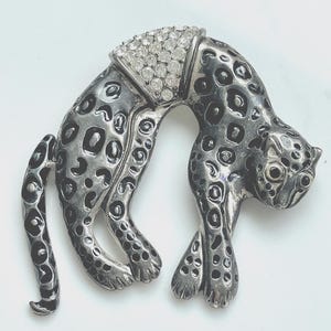May include: A silver-toned brooch in the shape of a leopard, with black spots and a curved pose. The leopard's head is adorned with a section of clear, sparkling crystals. The brooch is a decorative accessory.