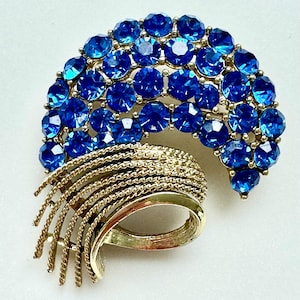 May include: A crescent-shaped brooch featuring numerous round, faceted blue rhinestones set in a gold-tone metal frame. The lower portion of the brooch consists of several curved, textured gold-tone metal ribbons that fan out in a layered design.