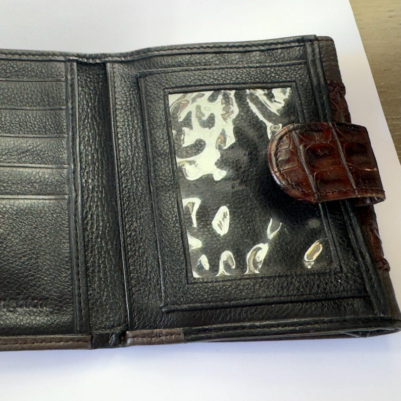 Puede incluir: Cartera de cuero negro abierta con un cierre marr&oacute;n con textura de cocodrilo. La cartera tiene m&uacute;ltiples ranuras para tarjetas y una ventana de identificaci&oacute;n transparente. El interior es de cuero negro con un acabado texturizado.