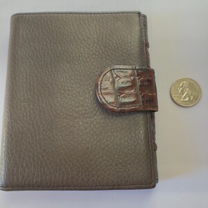 Puede incluir: Cartera de cuero gris con superficie texturizada y cierre marr&oacute;n con textura de cocodrilo. Una moneda est&aacute; al lado de la cartera para comparar el tama&ntilde;o. La cartera est&aacute; cerrada y es rectangular.