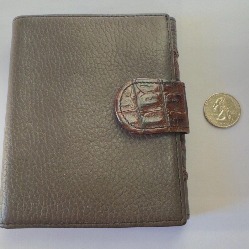 Puede incluir: Cartera de cuero gris con superficie texturizada y cierre marr&oacute;n con textura de cocodrilo. Una moneda est&aacute; al lado de la cartera para comparar el tama&ntilde;o. La cartera est&aacute; cerrada y es rectangular.