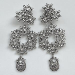 May include: A pair of silver-toned chandelier earrings. Each earring features a floral design with clear crystal accents. The earrings have a cluster of flowers at the top, a floral wreath in the middle, and a small, textured teardrop at the bottom.