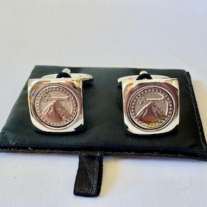 Op de afbeelding: Zilveren manchetknopen met het Paramount-logo, met een berg en de merknaam. De vierkante manchetknopen liggen op een zwart leren etui.