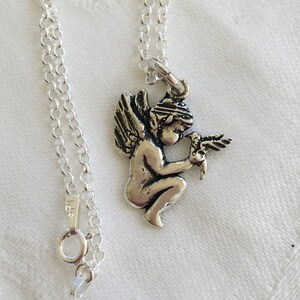 May include: A silver-colored necklace featuring a small pendant of a cherub holding a bird. The pendant is attached to a delicate chain with a lobster clasp. The necklace is displayed on a white surface.