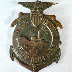 May include: A bronze-toned War Service Ship Building badge. The badge features an eagle at the top, a shield, and a ship illustration. The words "WAR SERVICE" and "SHIP BUILDING" are inscribed around the ship image.