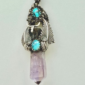 May include: A silver pendant featuring a bull silhouette, turquoise stones, and feather details. The pendant is attached to a purple amethyst crystal. The design incorporates floral accents and is suspended from a silver chain.