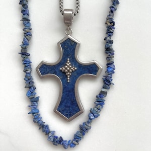 May include: A silver cross pendant with a dark blue inlay and a smaller silver cross design in the centre. A matching necklace of blue stone chips is also present, creating a coordinated jewellery set.