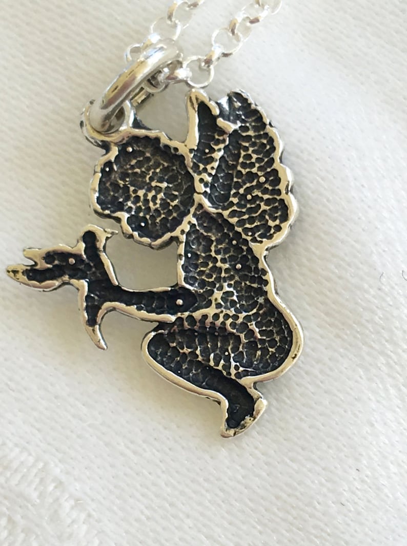 May include: A silver-toned pendant necklace featuring a detailed angel design. The angel is depicted in a seated pose, holding a sprig. The pendant has a textured, oxidized finish, and is suspended from a delicate chain.