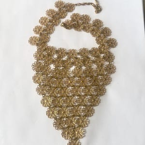 May include: A gold-toned necklace featuring a cascade of delicate, openwork floral designs. The necklace has a bib-style pendant and a chain with a clasp for adjustable length. The intricate details create a vintage aesthetic.