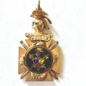 May include: A gold-colored pendant with a knight's helmet, bird, and cross design. The pendant features a black circular emblem with axes, a bird, and colorful letters. Two swords are crossed behind the emblem.