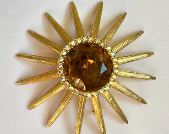 Peut inclure: Une broche en forme de soleil doré avec une grande pierre centrale facettée de couleur ambre. Le centre est entouré d'un anneau de petits strass clairs et étincelants. Les rayons du soleil présentent une finition en métal brossé texturé.