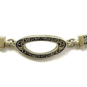 May include: A silver-toned bracelet featuring two textured, diagonal-patterned rectangular links connected to a central oval-shaped link with intricate dark detailing. The bracelet includes a toggle clasp with a T-bar and a circular ring closure.